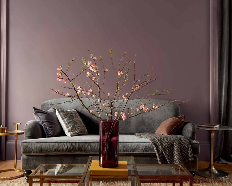A purple-painted living room features a gray couch, gold side tables, and a glass-topped gold coffee table topped with a red vase filled with pink flowers.